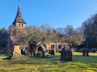Old brick church with a tall spire, trees, and gravestones under a clear blue sky.