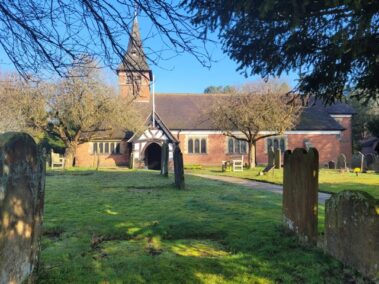 A brick church with a steeple, surrounded by gravestones and trees on a sunny day.