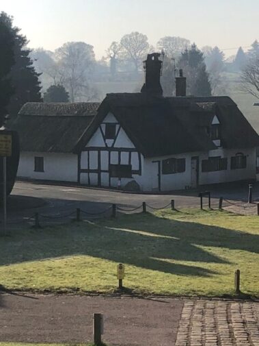 A quaint white cottage with a thatched roof stands beside a road on a misty morning.