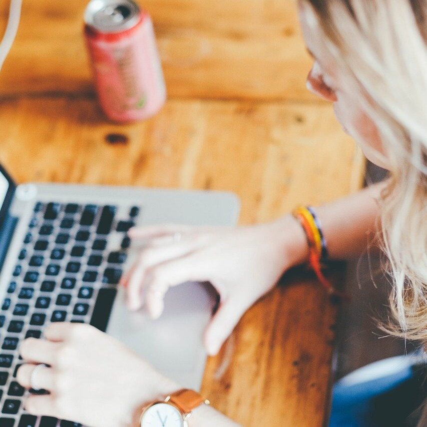 Person typing on a laptop at a wooden table with a drinks can nearby.