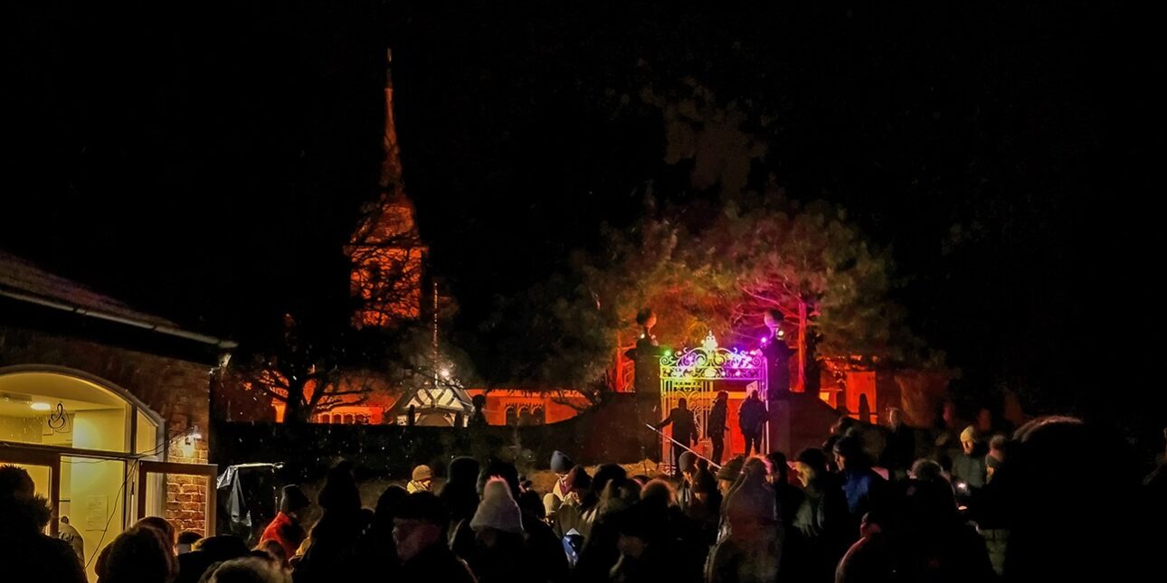 A crowd gathers at night near a colourful, lit stage and a church with an illuminated spire.