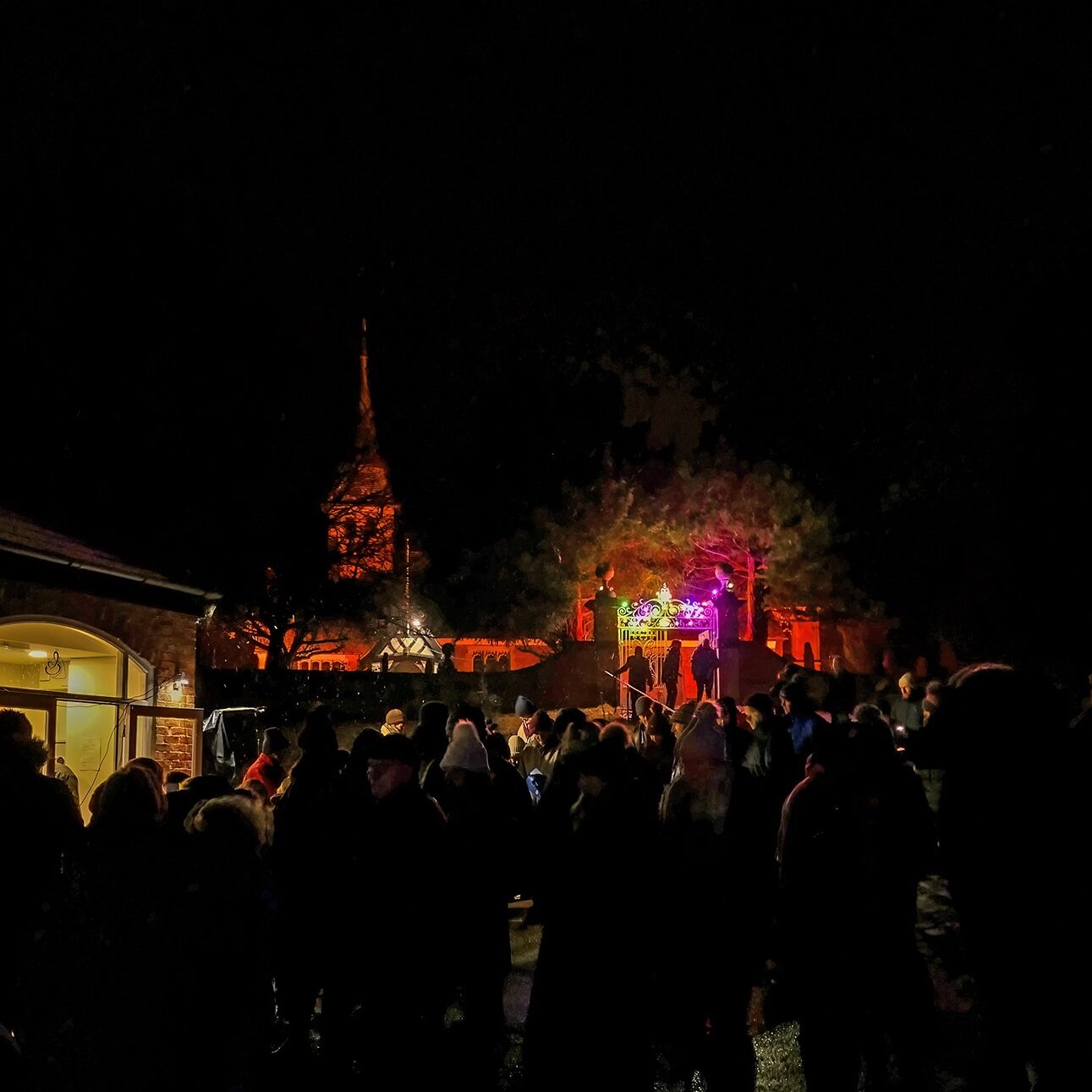 A crowd gathers at night near a colourful, lit stage and a church with an illuminated spire.