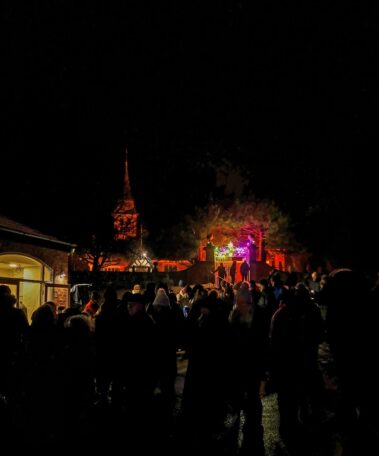 A crowd gathers at night near a colourful, lit stage and a church with an illuminated spire.