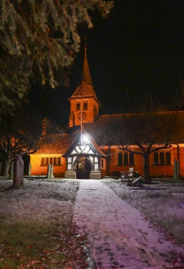 A church at night with a lit entrance, clock tower, and trees outlined against the dark sky.