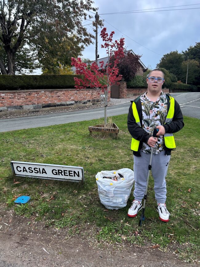 Person in yellow vest with litter picker stands by CASSIA GREEN sign and bag of rubbish.